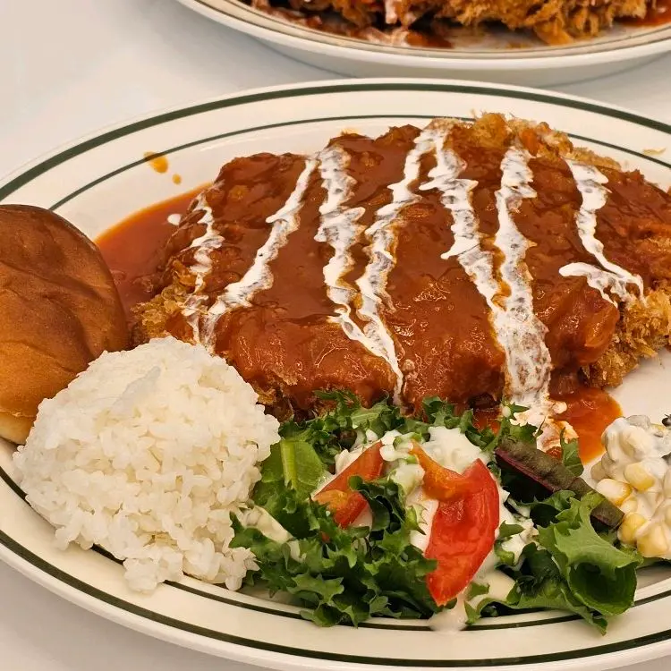 돈까스 위에 소스가 듬뿍 뿌려져 있고, 샐러드와 밥, 모닝빵이 함께 제공되는 모습