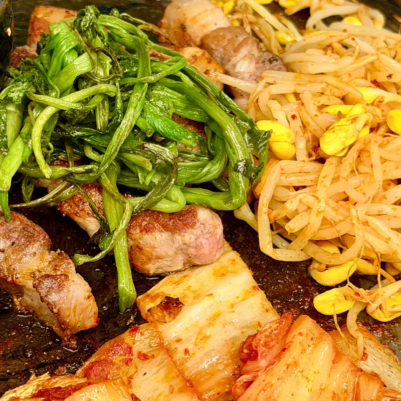 솥뚜껑 위에 구워진 삼겹살, 김치, 콩나물, 미나리의 푸짐한 모습