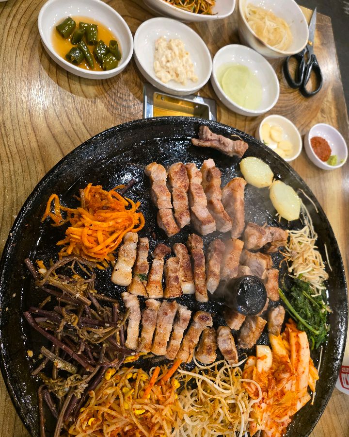 솥뚜껑 위에 삼겹살과 다양한 곁들임 채소가 함께 구워져 있는 모습