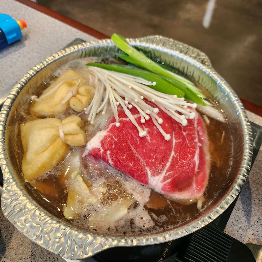 스키야키 냄비 안에는 신선한 야채와 얇게 썰린 소고기가 담겨 있다. 맑은 육수가 끓으면서 재료들의 풍미가 우러나오고 있다.