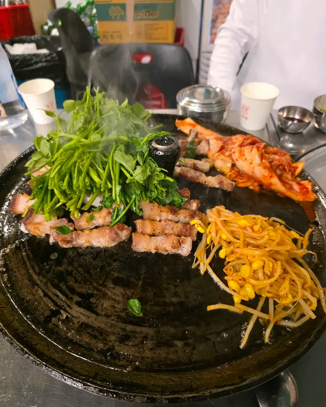 솥뚜껑 위에 삼겹살, 김치, 콩나물, 미나리가 함께 구워지고 있는 모습