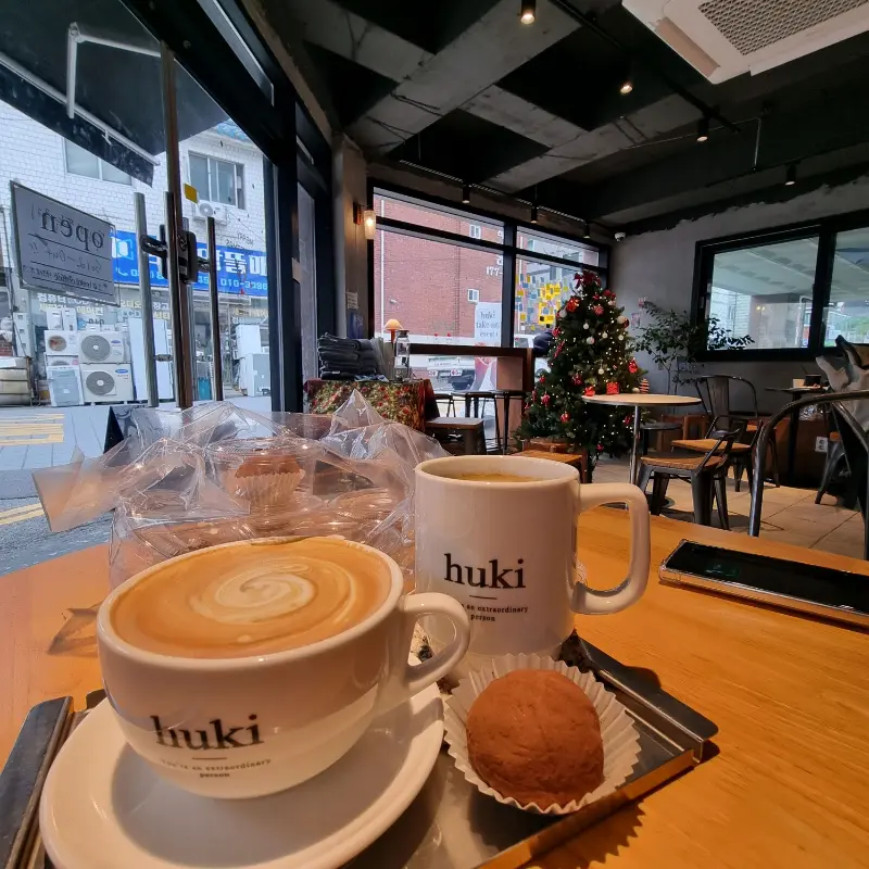 따뜻한 라떼와 디저트가 테이블 위에 놓여있는 모습. 'huki' 로고가 새겨진 컵이 인상적이다.