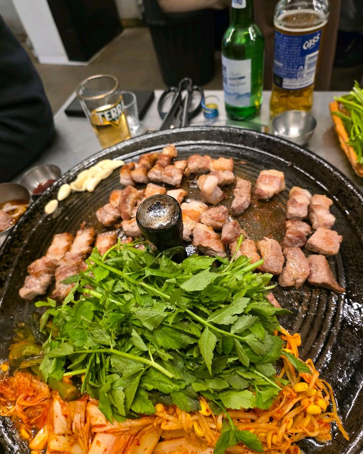 솥뚜껑에 구워진 삼겹살, 김치, 콩나물, 미나리