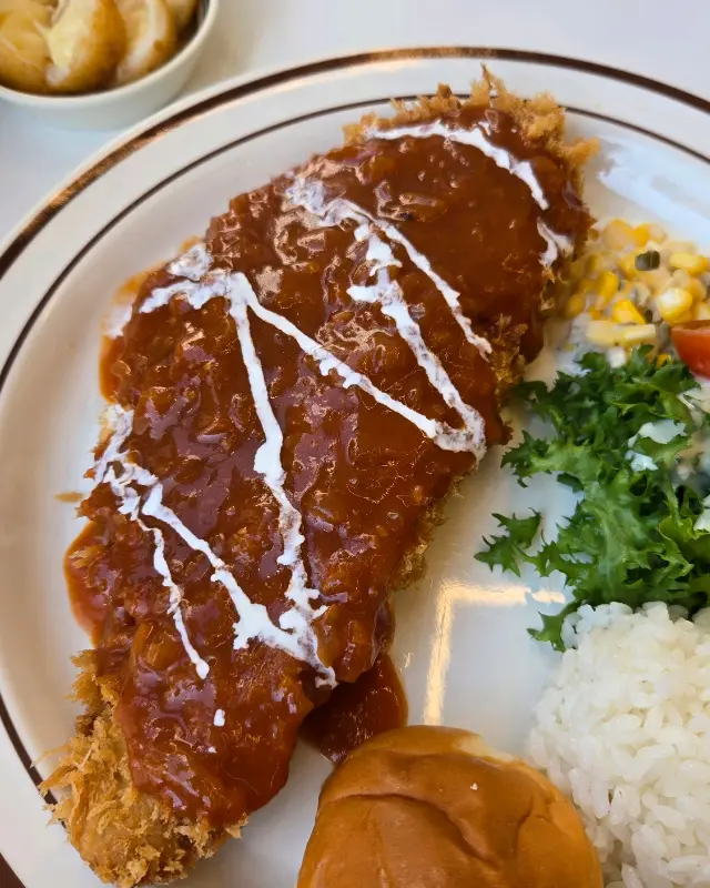 돈까스 위에 소스와 하얀색 소스가 뿌려져 있고, 샐러드와 밥, 모닝빵이 함께 제공되는 모습