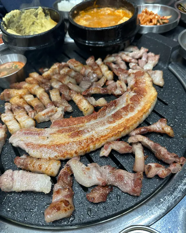 불판 위에 삼겹살이 올려져 있고, 주변에 청국장과 계란찜 등 다양한 밑반찬이 놓여 있는 모습