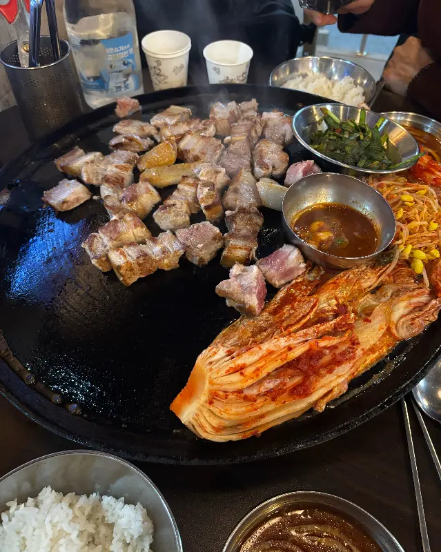 솥뚜껑 위에서 맛있게 익어가는 삼겹살과 곁들임, 밥그릇과 소스 그릇