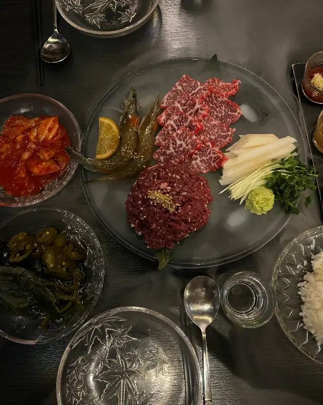 뭉티기, 육회, 간장새우가 한 접시에 담겨 있고, 김치와 소스, 밥 등의 밑반찬이 테이블 가득 차려진 모습