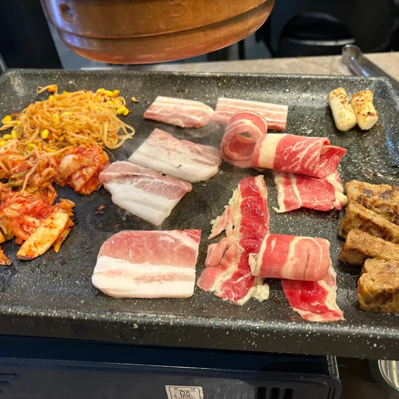 불판에 구워지는 고기, 김치, 콩나물, 떡볶이 떡 사진