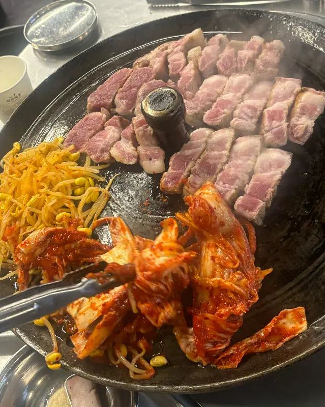 솥뚜껑 위에 삼겹살, 김치, 콩나물이 함께 구워지고 있는 모습