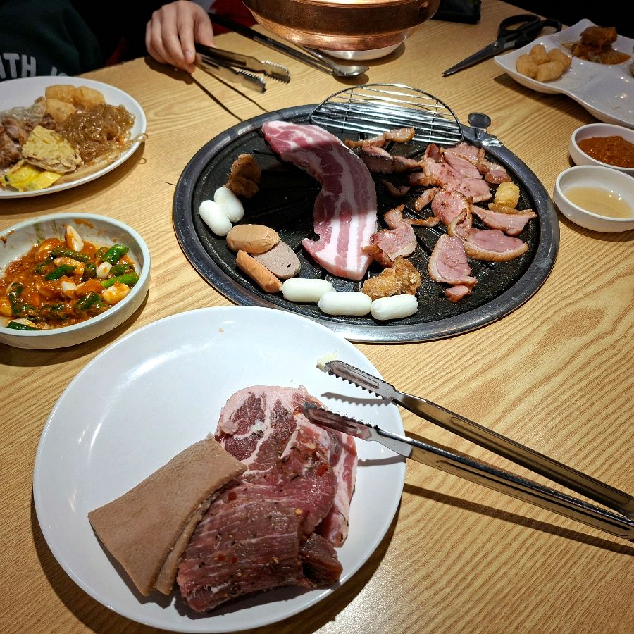 불판 위에 삼겹살, 닭갈비, 소시지, 떡 등이 함께 구워지고 있는 모습