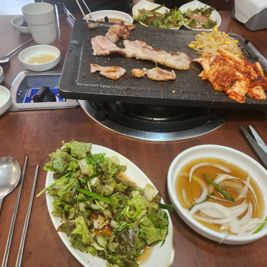 불판 위에 삼겹살과 김치, 콩나물을 올려 구워 먹는 모습. 다양한 밑반찬도 눈에 띕니다.