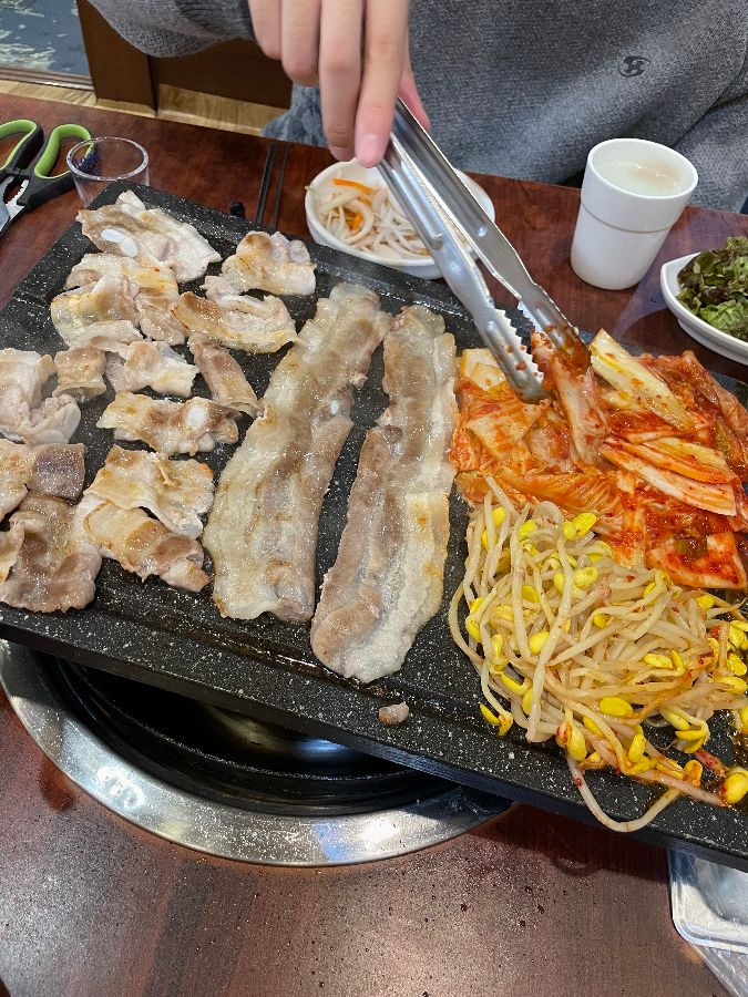 노릇하게 구워진 삼겹살을 먹기 좋은 크기로 자르는 모습. 김치와 콩나물도 함께 구워져 풍성함을 더합니다.