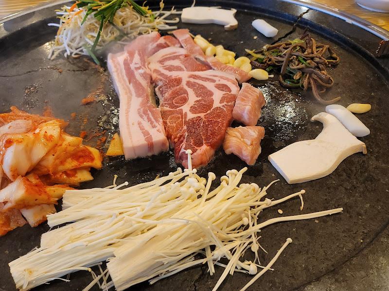철판 위에 삼겹살, 김치, 콩나물, 버섯, 마늘 등이 함께 구워지고 있는 모습
