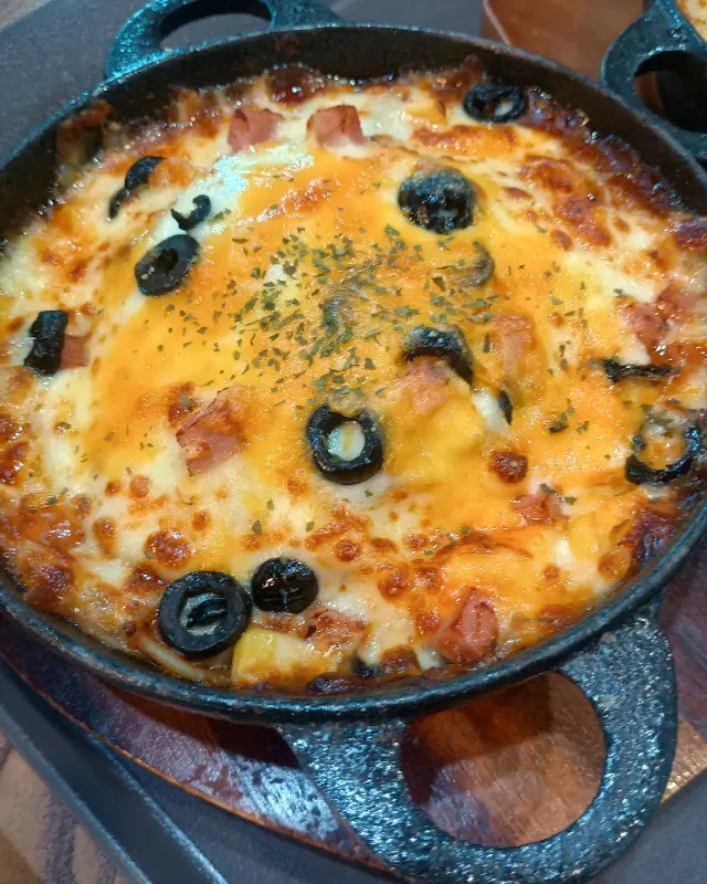 고구마 무스 피자의 클로즈업 샷. 검은색 주물 팬에 담겨져 있으며, 치즈가 녹아내리고 검은 올리브가 토핑으로 올려져 있다.