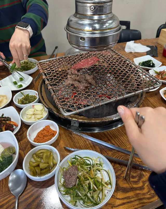 숯불 위에 석쇠가 놓여 있고, 그 위에 불고기가 구워지고 있으며, 다양한 밑반찬 그릇들이 테이블을 가득 채우고 있는 모습