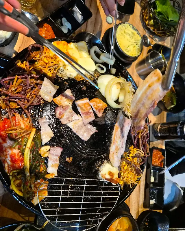 솥뚜껑 위에 삼겹살, 김치, 콩나물, 고사리, 파김치 등이 푸짐하게 구워지고 있는 모습