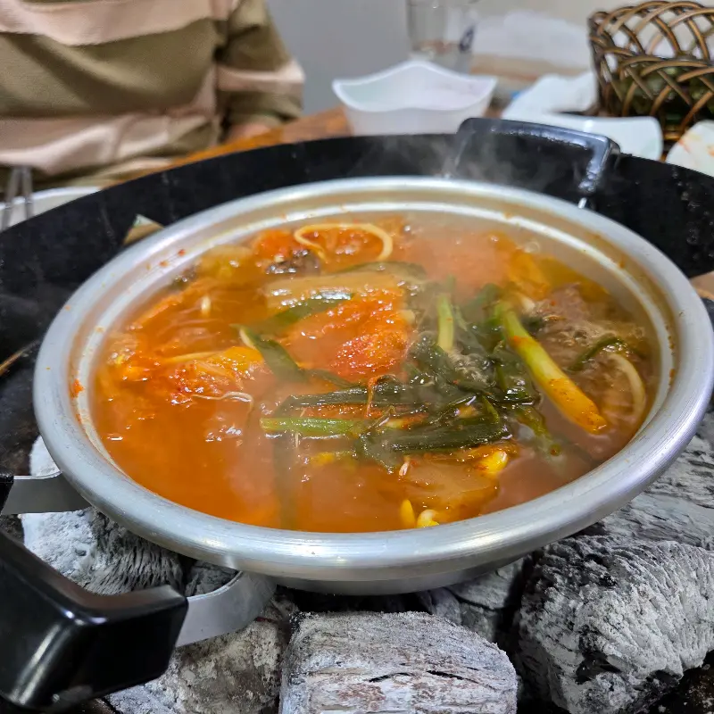 숯불 위에 올려진 뚝배기 안에는 김치와 다양한 채소가 듬뿍 들어간 김치국이 담겨져 있고, 김이 모락모락 피어오르는 모습