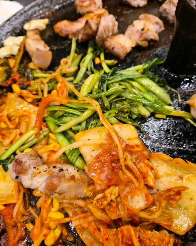 솥뚜껑에 구워진 삼겹살, 김치, 콩나물, 미나리의 모습