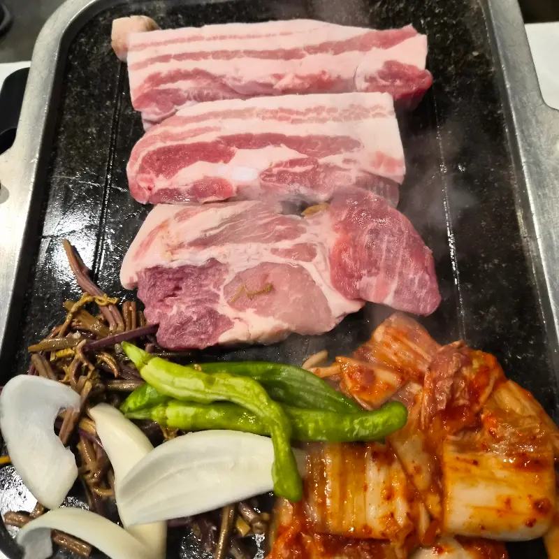 불판 위에 가지런히 놓인 삼겹살과 김치, 고사리, 양파