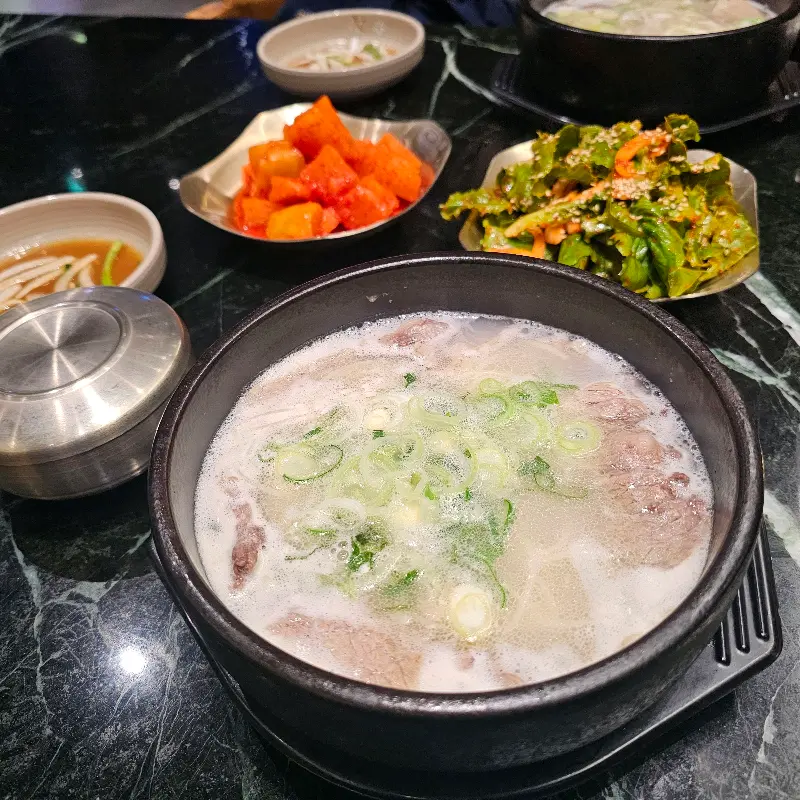 테이블 위에 설렁탕과 깍두기, 겉절이 등이 놓여있고, 뚝배기 안에는 설렁탕이 담겨 있다.