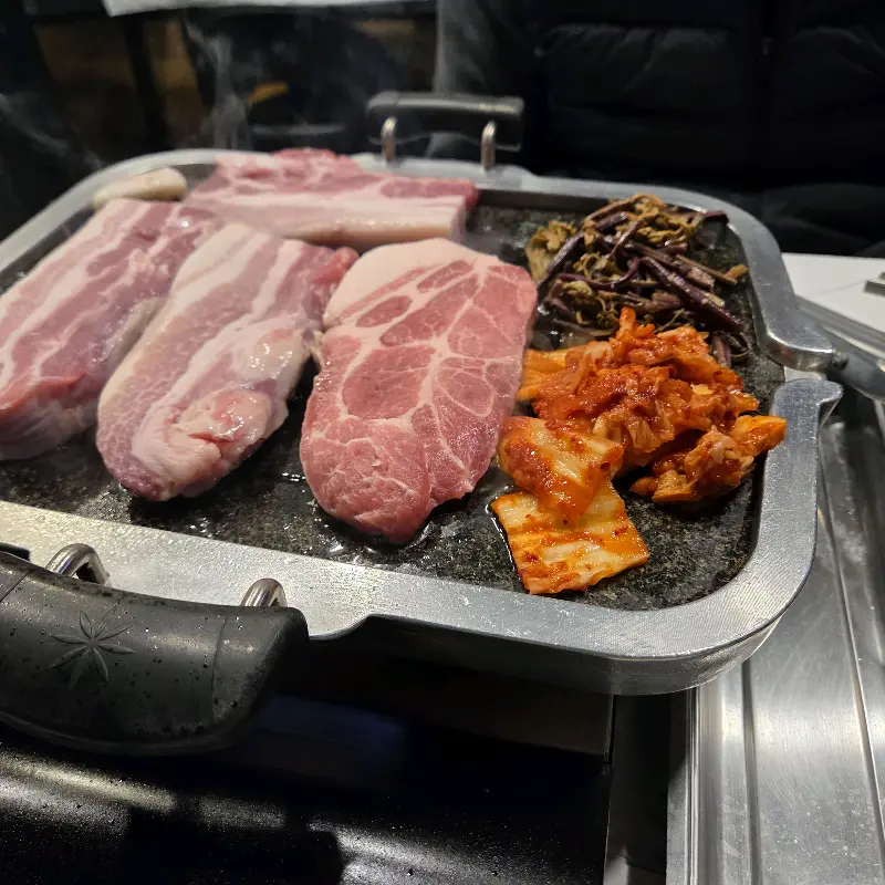 불판 위에 올려진 삼겹살, 김치, 고사리