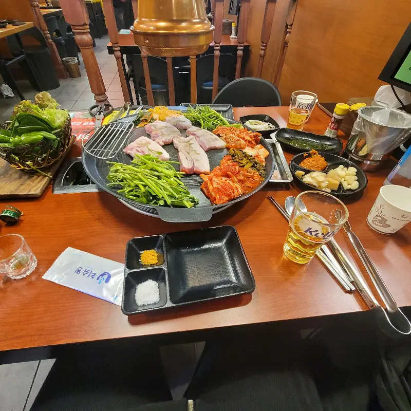 테이블 위에 솥뚜껑, 삼겹살, 각종 반찬들이 푸짐하게 차려진 모습