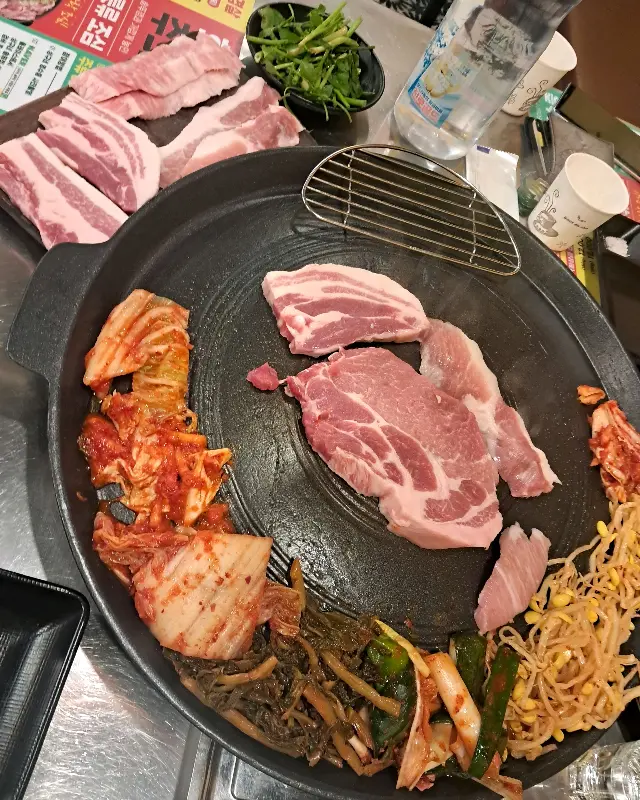 솥뚜껑 위에 삼겹살, 김치, 콩나물, 시래기 등이 함께 구워지고 있는 모습