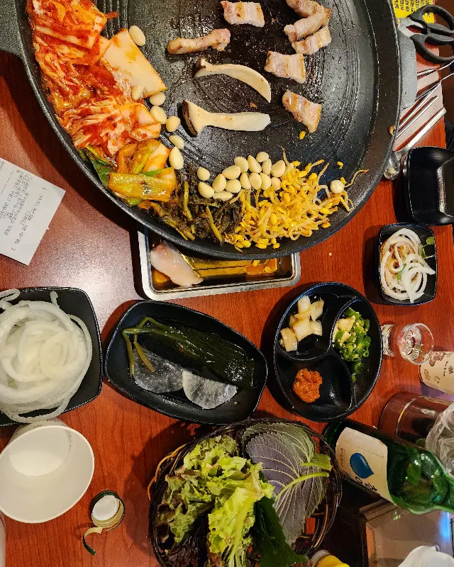 솥뚜껑 위에서 삼겹살, 김치, 콩나물, 버섯 등이 함께 구워지고 있는 모습