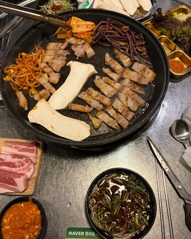 불판 위에 삼겹살, 김치, 콩나물, 버섯이 함께 구워지는 모습