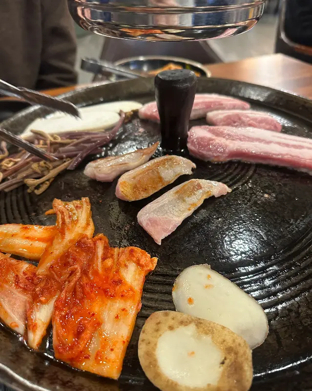 솥뚜껑에 구워진 삼겹살, 김치, 감자의 먹음직스러운 모습