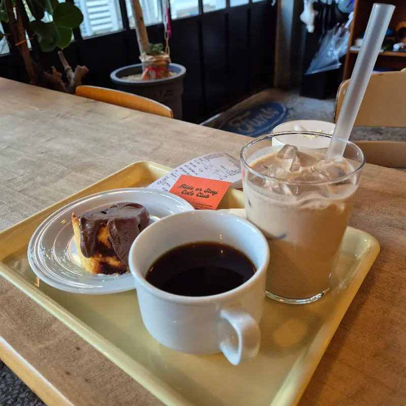 테이블 위에 놓인 커피와 케이크. 햇살이 들어오는 창가 자리에 앉아 커피와 케이크를 즐기는 모습.