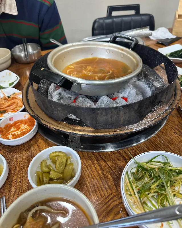 숯불 화로 위에 김치국이 담긴 냄비가 올려져 있고, 주변에는 다양한 밑반찬 그릇들이 놓여 있는 모습