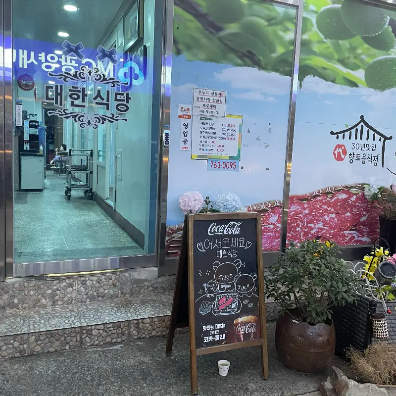 대한식당 입구에는 메뉴판과 코카콜라 곰돌이 그림이 그려진 입간판이 세워져 있고, '어서오세요 대한식당'이라는 문구가 쓰여 있는 모습