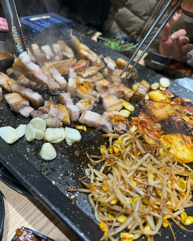 불판 위에서 지글거리는 삼겹살, 마늘, 콩나물, 김치