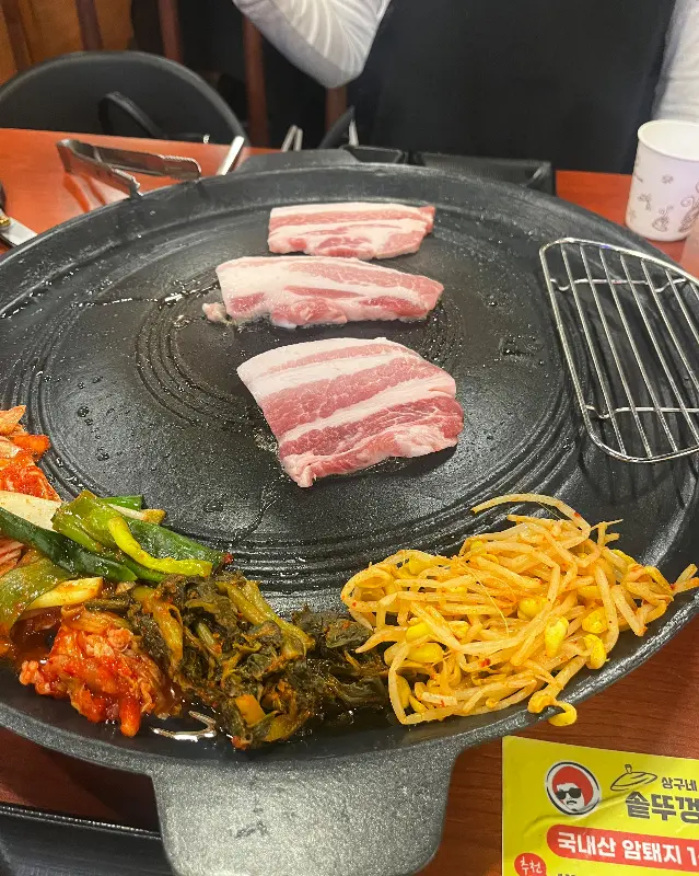 솥뚜껑에 삼겹살, 김치, 콩나물 등이 구워지고 있는 모습