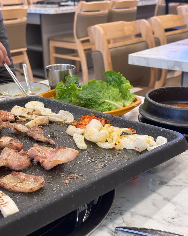 잘 구워진 삼겹살과 버섯, 마늘이 불판 위에 올려져 있고, 싱싱한 상추쌈이 옆에 놓여 있다.
