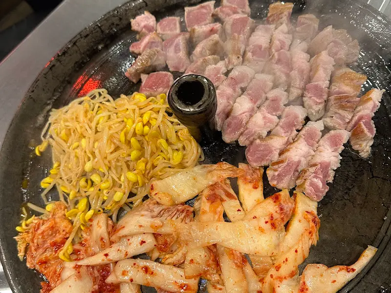 솥뚜껑 위에 삼겹살, 김치, 콩나물이 함께 구워지고 있는 모습