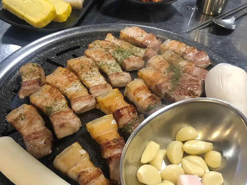 불판 위에 구워진 오겹살, 버섯, 떡, 마늘이 함께 놓여 있다.