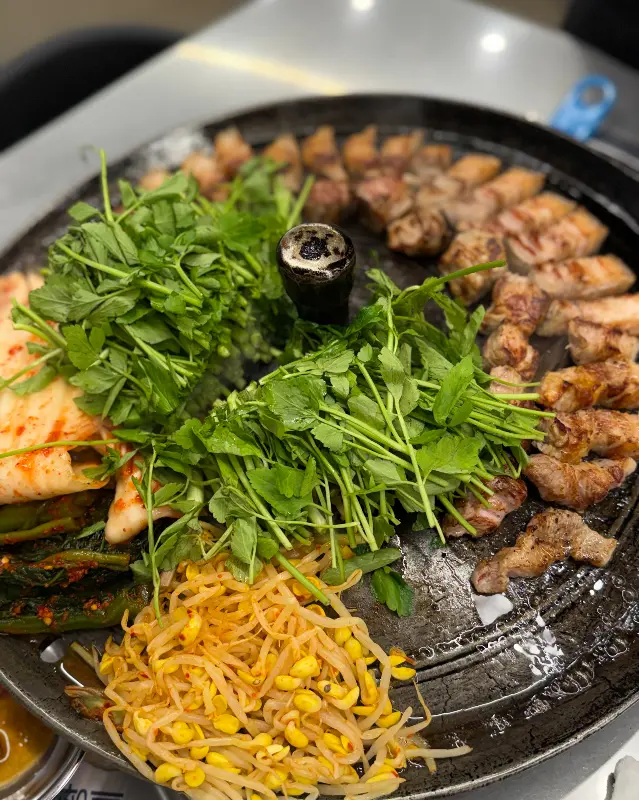 솥뚜껑 위에 올려진 잘 익은 고기, 김치, 미나리