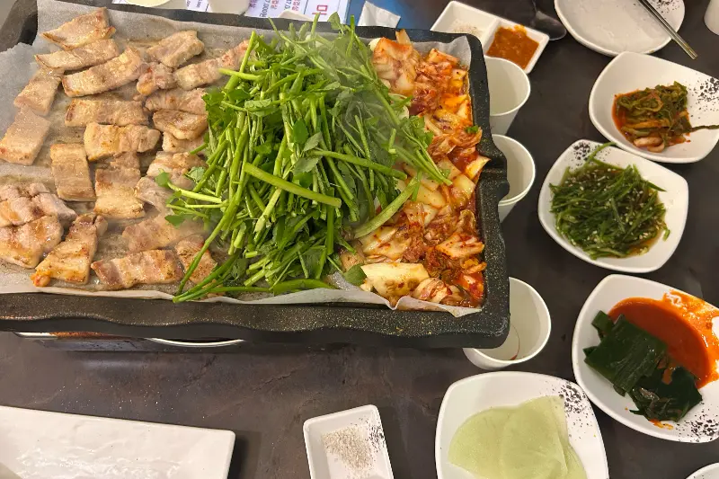 불판 위에 삼겹살, 미나리, 김치가 함께 구워지는 모습