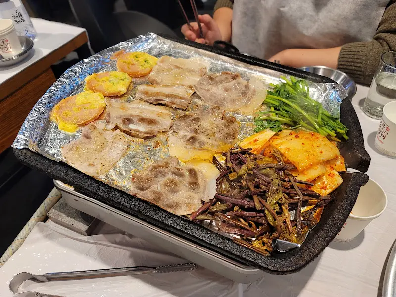 불판 위에 냉동 삼겹살, 김치, 고사리, 미나리가 함께 올려져 있는 모습