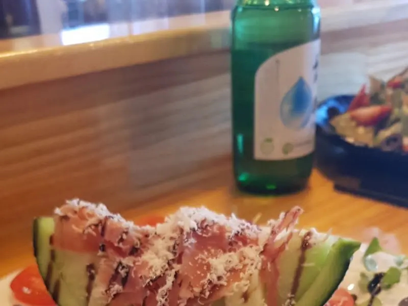 멜론 위에 햄과 치즈가 올려진 요리가 놓여 있고, 그 뒤로 물병과 샐러드가 보인다.