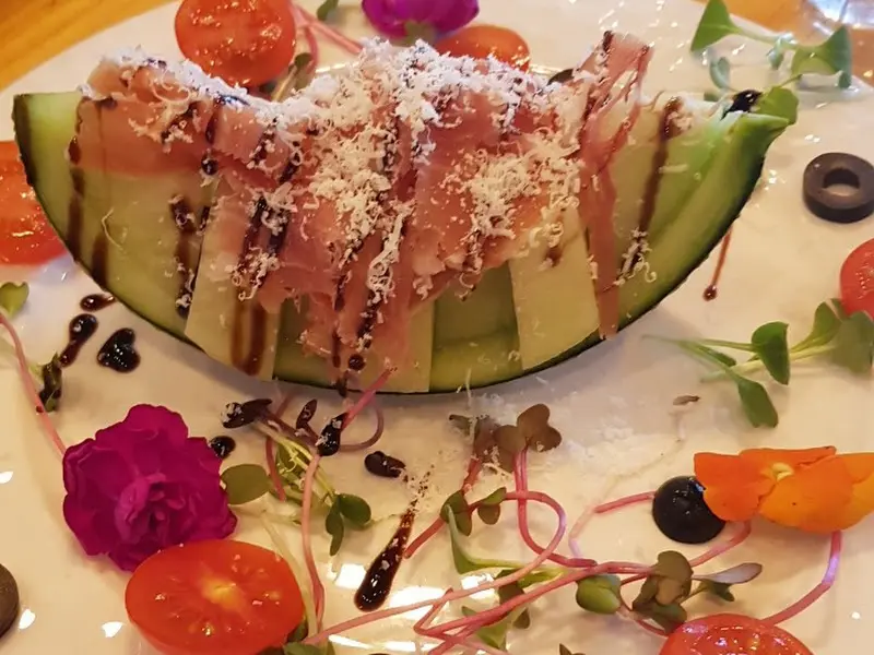 멜론 위에 얇게 저민 햄이 올려져 있고, 그 위에 하얀 치즈가 눈처럼 뿌려져 있다. 접시 주변에는 붉은색, 주황색, 보라색의 식용 꽃과 채소가 장식되어 있다.
