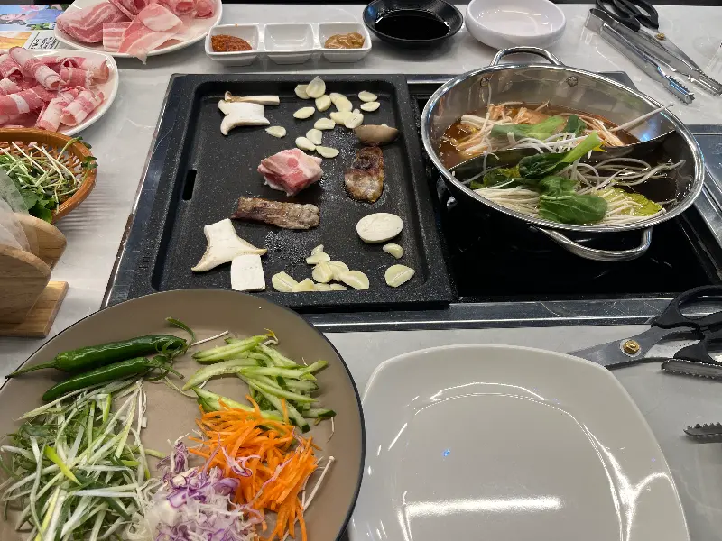 테이블 위에 놓인 샤브샤브 냄비와 고기 불판, 그리고 다양한 채소와 소스