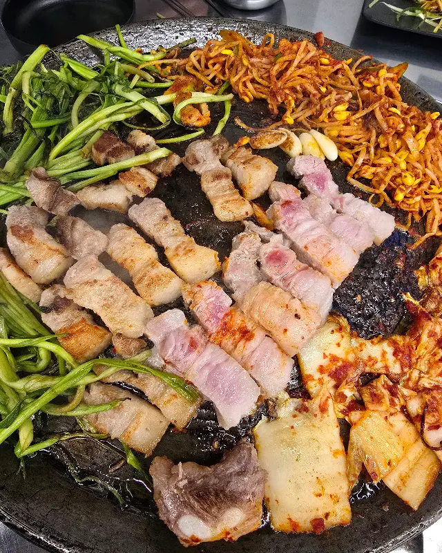 노릇하게 구워진 삼겹살과 김치, 콩나물, 미나리의 모습