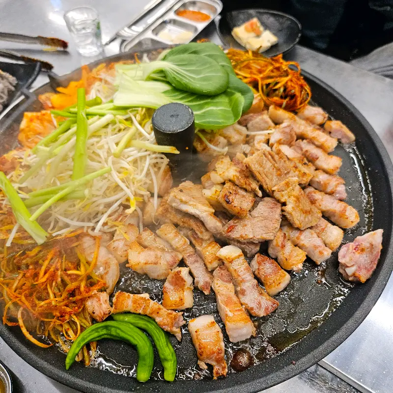 솥뚜껑 위에 삼겹살과 다양한 야채, 김치가 푸짐하게 구워져 있는 모습