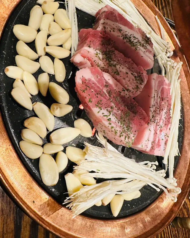 불판 위에 두툼한 삼겹살과 마늘, 버섯이 올려져 있는 모습