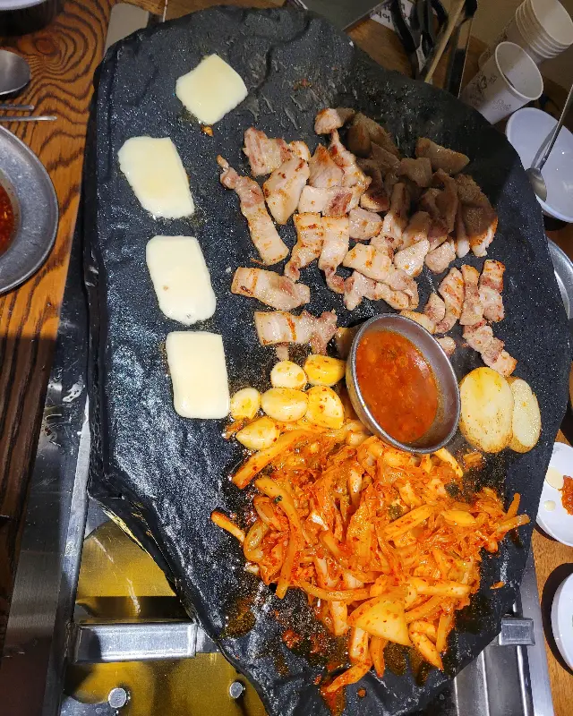 돌판 위에 구워진 삼겹살, 김치, 치즈, 마늘