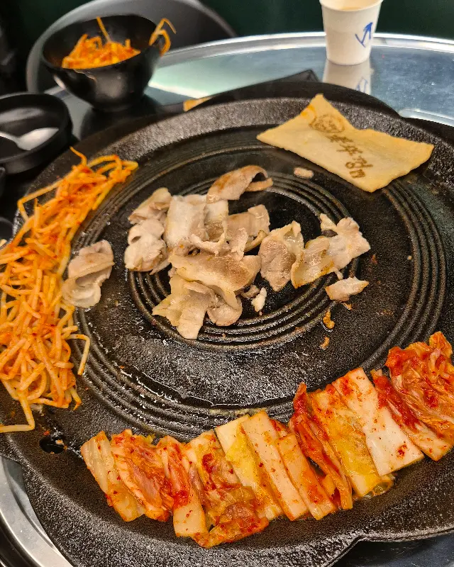 불판 위에 삼겹살과 김치가 맛있게 구워져 있고, 옆에는 시원한 물쫄면이 놓여 있는 모습