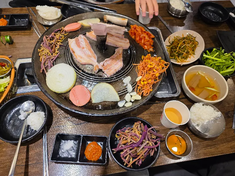 솥뚜껑 위에 삼겹살, 김치, 콩나물, 고사리, 미나리, 양파, 버섯, 소시지까지 푸짐하게 구워진 모습